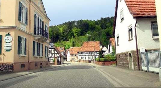 Dorfstraße mit Fachwerkhäusern, beigem Gebäude links, weißem Haus rechts und bewaldetem Hügel im Hintergrund.