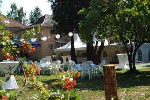 Gartenfest mit weißen Pagodenzelten, Tischen und Plastikstühlen unter Bäumen, Lichterketten und roten Beeren im Vordergrund.