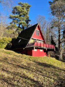 Rotes A-förmiges Ferienhaus mit Balkon in bewaldeter Hanglage unter blauem Himmel
