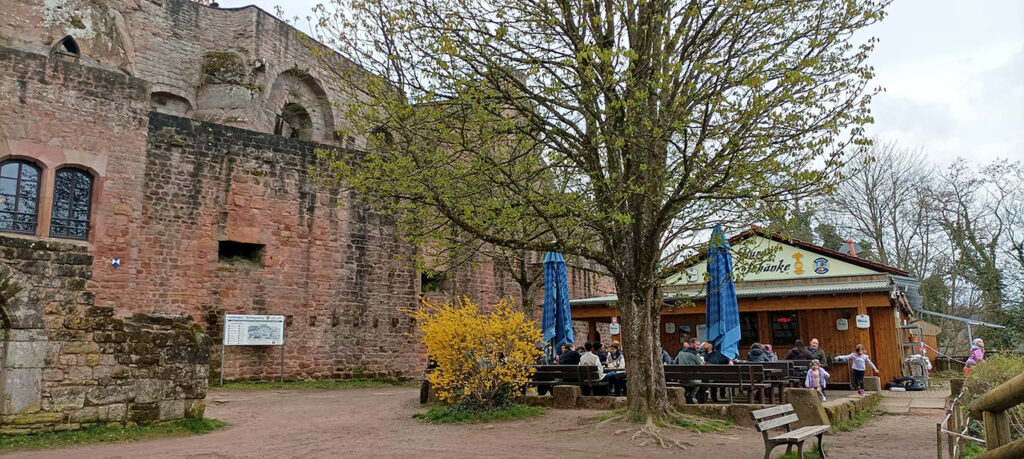 Blick auf eine große Steinburg mit einer Außenbereichsgaststätte, Tische, Bänke, blauer Sonnenschirm und Menschen neben einem Baum.