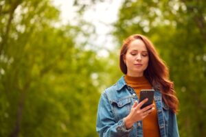 Frau mit roten Haaren und Jeansjacke schaut auf ihr Smartphone in einem grünen Park.