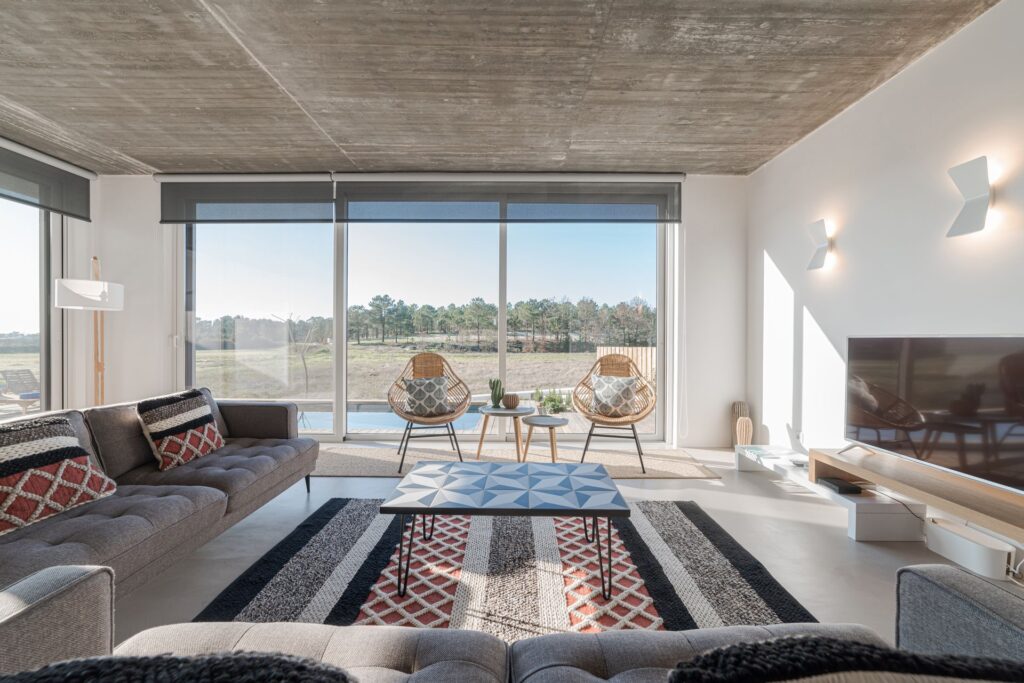 Modernes Wohnzimmer mit großen Fenstern, grauen Sofas, gemustertem Teppich und zwei geflochtenen Stühlen vor Naturlandschaft.