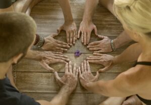 Mehrere Hände liegen im Kreis auf einem gealterten Holztisch, um eine kleine violette Blüte in der Bildmitte gelegt.