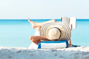 Person liegt am Strand auf einem Handtuch, trägt großen gestreiften Sonnenhut und liest vor türkisblauem Meer.