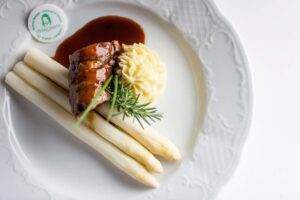 Drei weiße Spargelstangen mit einer Scheibe glasierter Fleischroulade, Kartoffelpurée und Rosmarienzweig auf einem dekorativen weißen Teller.