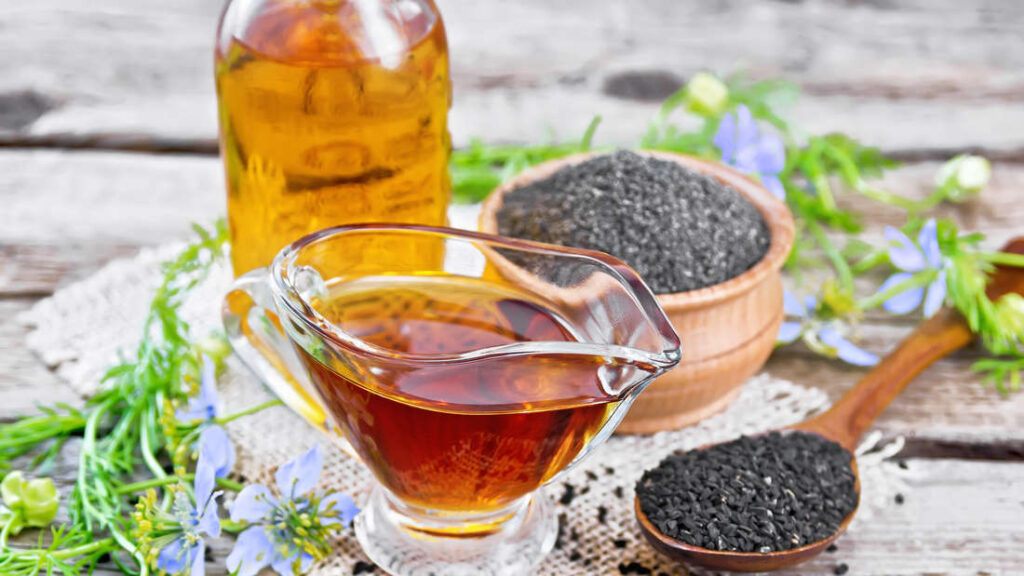 Glasgießer mit bernsteinfarbenem Schwarzkümmelöl, im Hintergrund Holzlöffel und Schale mit schwarzen Kümmelsamen und blauen Blüten auf Holzplatte