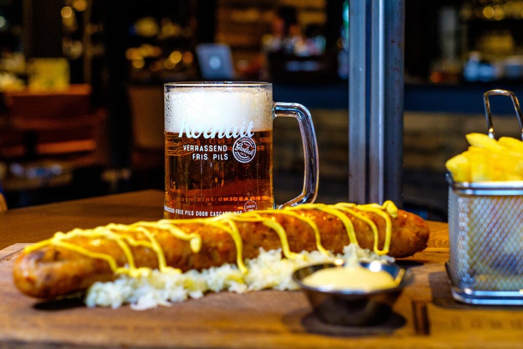 Großes Bierglas mit Schaum neben einer länglichen Bratwurst mit Senf und Pommes in einem Restaurantambiente