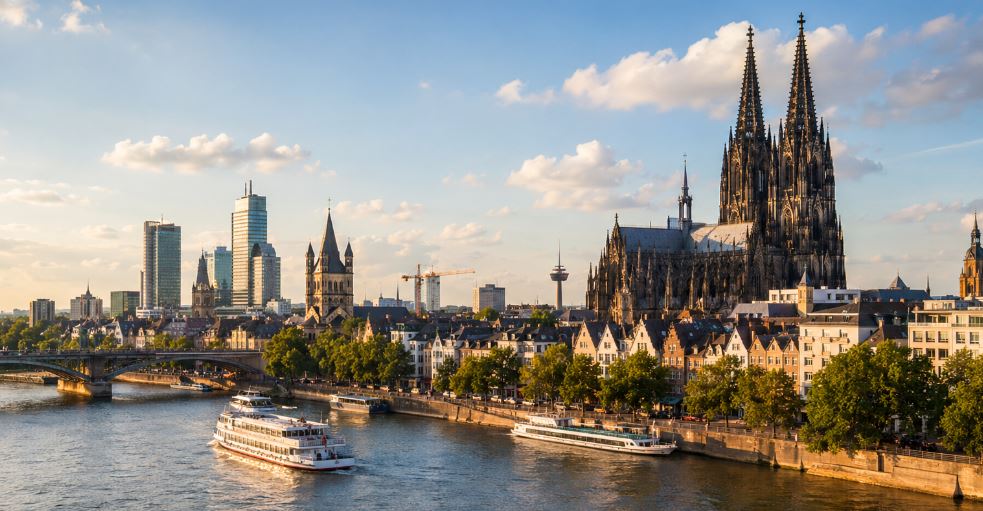 Kölner Dom und Altstadt an einem sonnigen Flussufer mit Ausflugsbooten auf dem Rhein und moderner Skyline im Hintergrund.