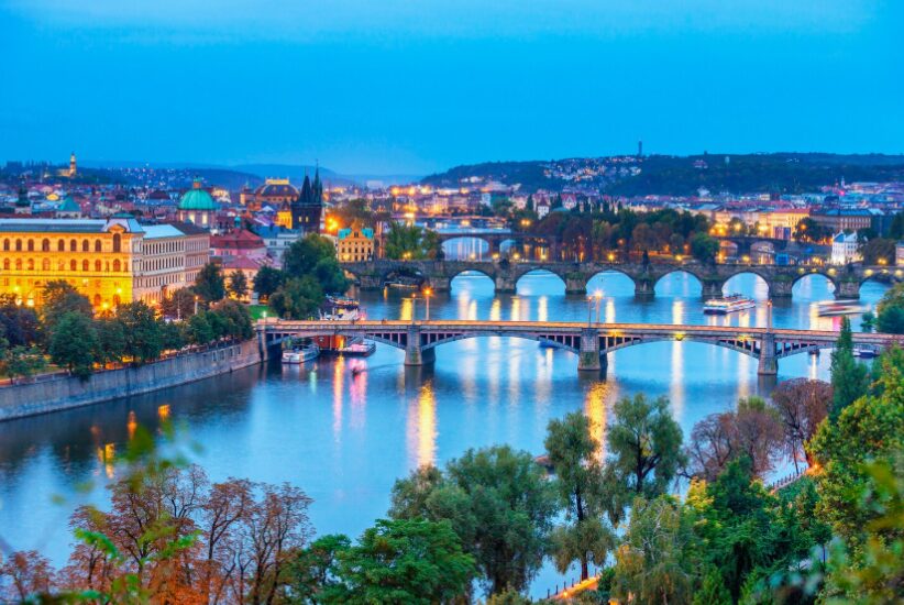 Blick über die Moldau in Prag bei Dämmerung mit mehreren beleuchteten Brücken und Ufergebäuden, reflektierende Lichter im Wasser