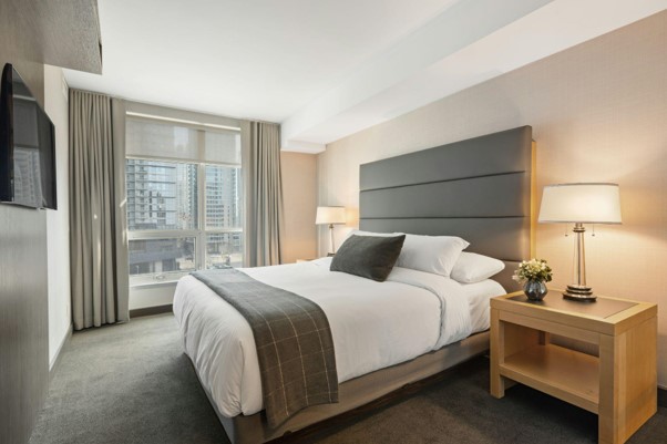 Helles Hotelzimmer mit großem Bett, gepolstertem Kopfteil, Nachttisch mit Lampe und Fenster mit Stadtblick.