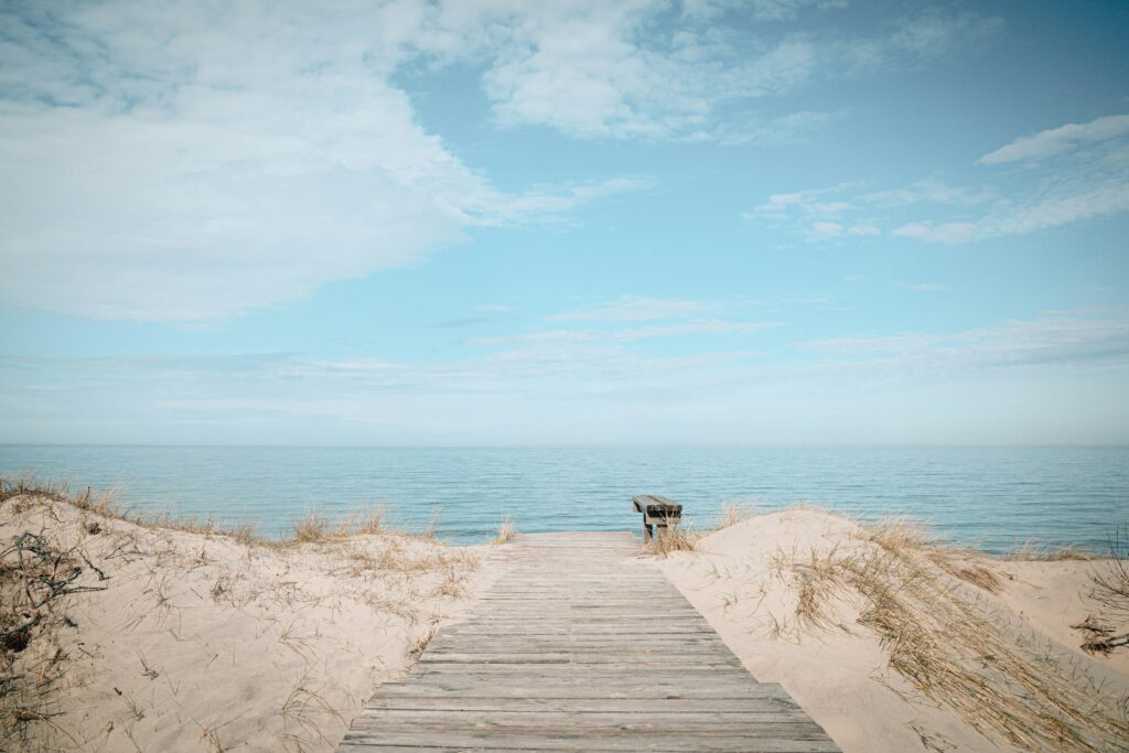 Holzsteg führt durch Sanddünen zum ruhigen Meer unter blauem Himmel mit Wolken
