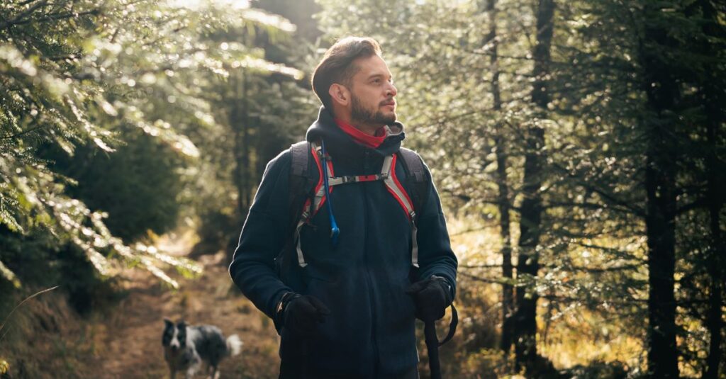 Wanderer mit Rucksack und Wanderstock auf Waldweg, im Hintergrund ein Hund
