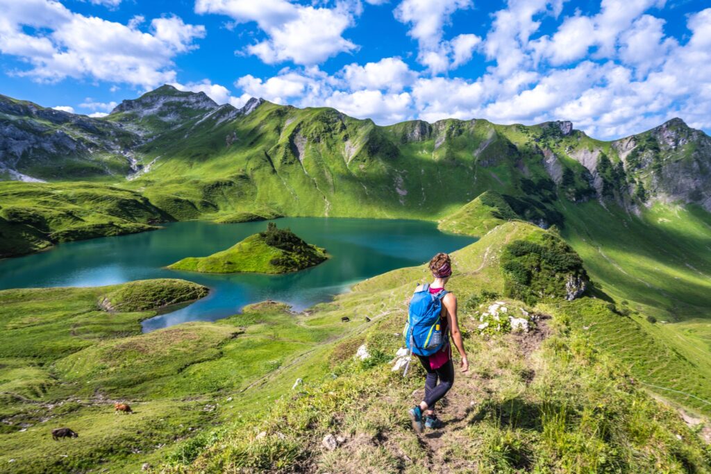 Wanderin blickt über einen Bergsee in grüner Alpenlandschaft mit steilen Gipfeln und blauem Himmel